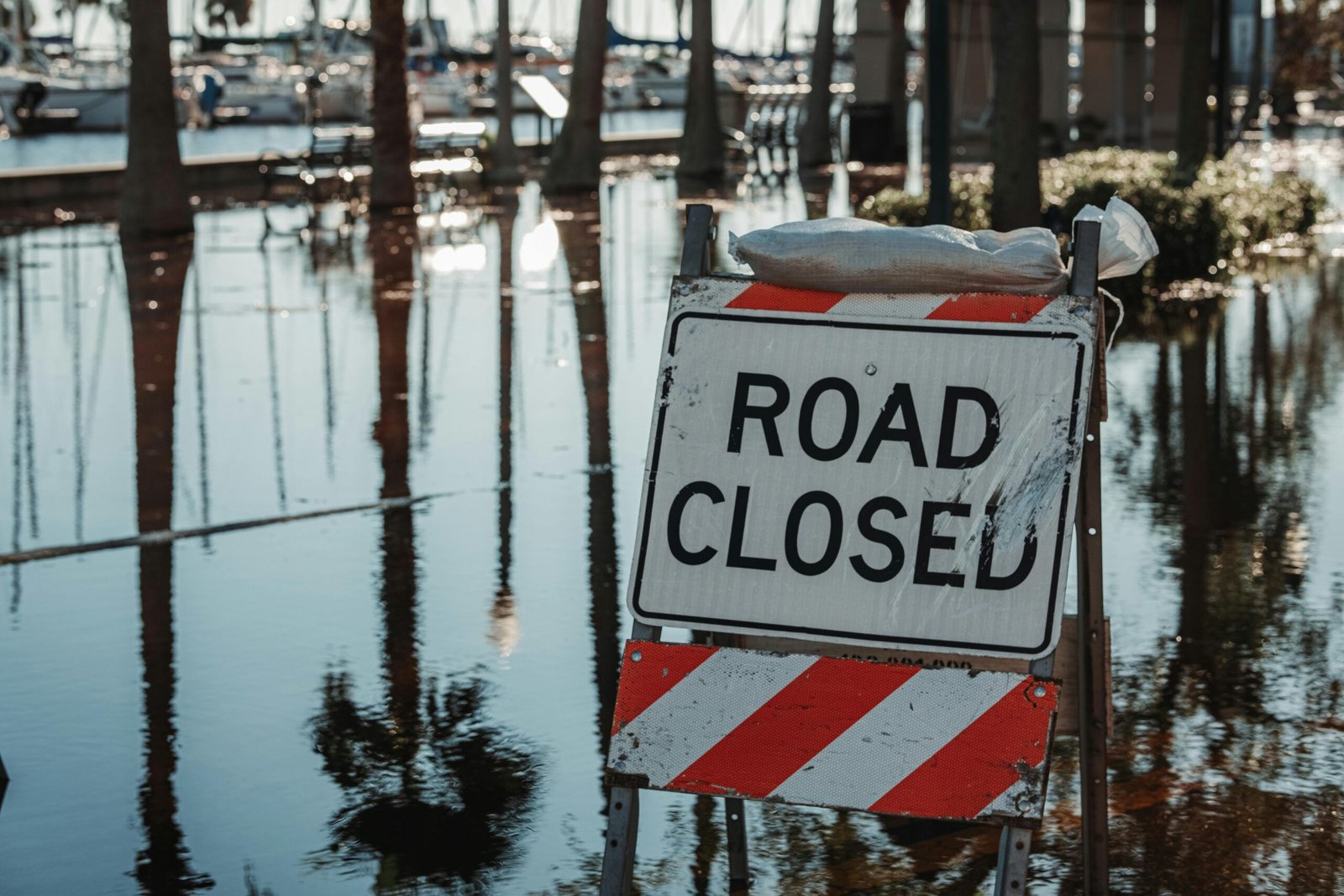 Road closed sign amidst flooded street, reflecting calm water and trees.